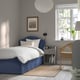 Blue curved-headboard bed, white linens, grey bedside table, plant, wicker chair, brown rug.