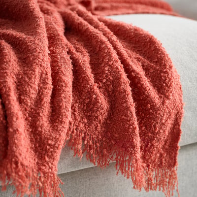 Bright orange-red textured throw blanket draped over light-colored cushion. Soft, looped yarn creates an uneven, lustrous surface.