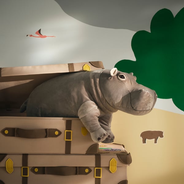 A whimsical grey hippo toy sits atop two brown trunks with gold buckles against a wall with green leaf and pink flamingo stickers.