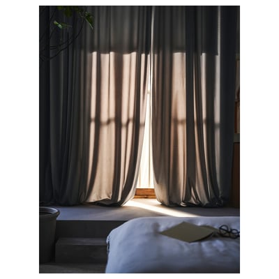 Soft, dark grey curtains partially open, letting sunlight gently philtre through, revealing a serene bedroom scene with a bed and personal items.