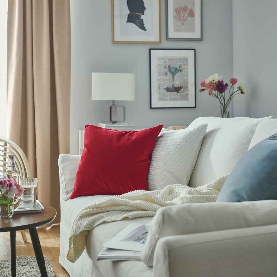 Modern living room with white sofa, red SANELA cushion, table lamp, and flower vase on wooden table, blue curtains.