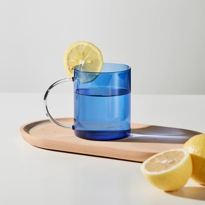 Blue mug with curved handle on tray, lemon slice on rim and half lemon beside, suggests lemony drink.