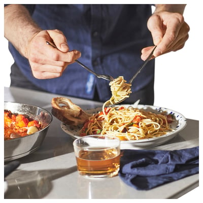 A chef twirls spaghetti with tongs over a plate. Next to it, a glass of beer and a bowl of salad.