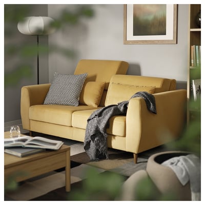 Modern yellow sofa with grey pillows, soft fabric, sleek design, and wooden legs near a coffee table with books and a glass.