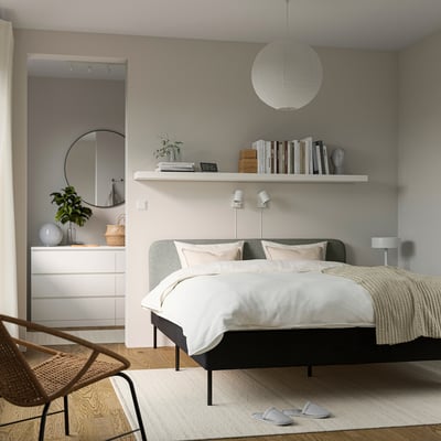 Minimalist bedroom with white bed, grey headboard cover, hanging light, bookshelf, mirror, dresser, and a wicker chair. White slippers on rug.