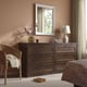 Wooden dresser w/mirror, lamp, flowers. Dark brown, 6 drawers. Wicker chair with blanket. Bed to right.