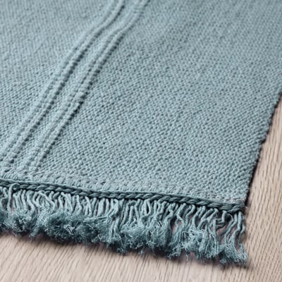 Handcrafted light blue rug with fringes, placed on wooden floor.