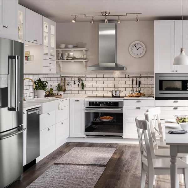 Modern white kitchen with stainless steel appliances, wooden floors, and ample lighting. Includes oven, microwave, dishwasher, and large wall clock.