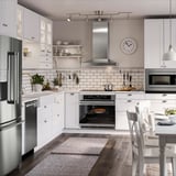 Modern white kitchen with stainless steel appliances, wooden floors, and ample lighting. Includes oven, microwave, dishwasher, and large wall clock.