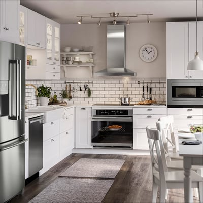 Modern white kitchen with stainless steel appliances, wooden floors, and ample lighting. Includes oven, microwave, dishwasher, and large wall clock.
