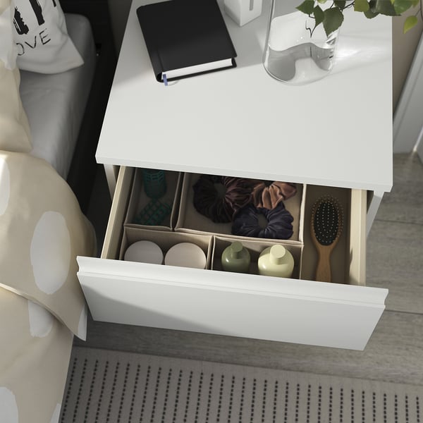 STORKLINTA white bedside table, modern design, smooth drawers, integrated handles, organised with small items, brush, and bottles.