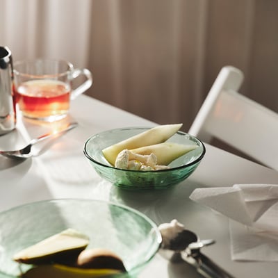 Bowl of sliced melon on a table, accompanied by a glass of tea, showcasing the STRANDKRABBA bowls inviting shape and practical stacking feature.