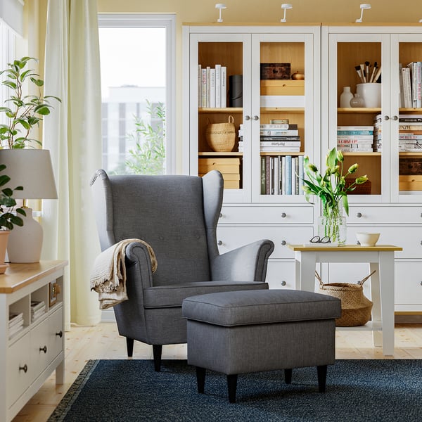 Cosy grey STRANDMON chair & ottoman in living room. Comfortable, upholstered, & large, set beside tall white bookshelves.