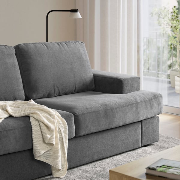 A grey SUNDBRON sofa sits near a window. Its modern design fits well in a cosy living room. A cream blanket curtains over the arm.