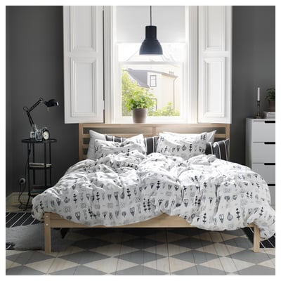 A cosy bedroom with a light-colored wooden TARVA bed frame, white linens, and black accents. A large window reveals a house outside.