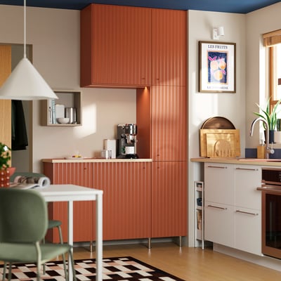 Modern kitchen with terracotta-colored cabinets, white appliances, green chair, checkerboard rug, and fruit poster.