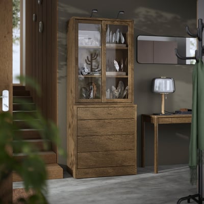 Smoked oak TONSTAD cabinet & chest with brass handles & glass doors, housing decor, taller than console table & lamp, adding warmth & function.
