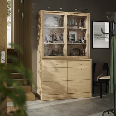 Light wooden TONSTAD cabinet with drawers and glass doors stores decorated items. Located next to stairs and chair, provides ample storage.