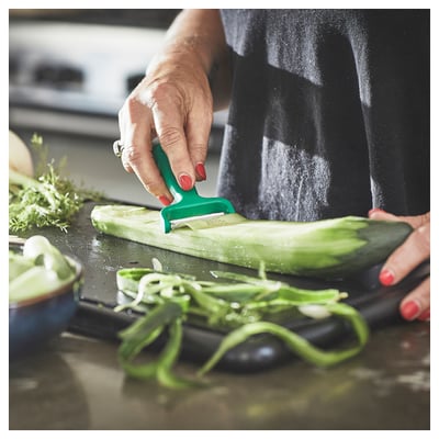 A person uses a green UPPFYLLD peeler on a cucumber, creating vegetable strips on a cutting board.