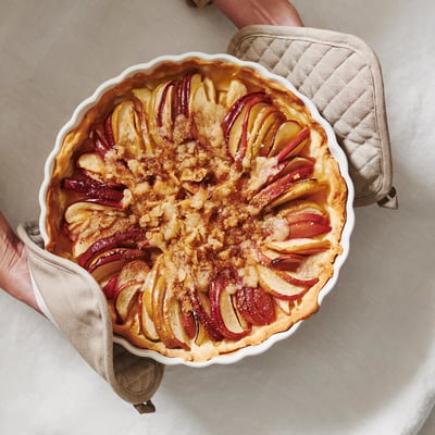 A person holding a freshly baked apple pie in a beige dish, showing the quilted potholders for heat protection.