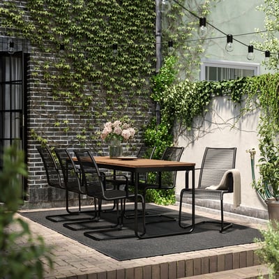 Modern outdoor dining setup with black metal VÄSMAN table and chairs, surrounded by greenery and string lights.