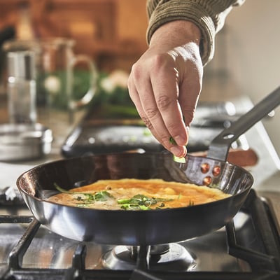 A hand stirs a fried dish with herbs in a black carbon-steel frying pan on a stove.