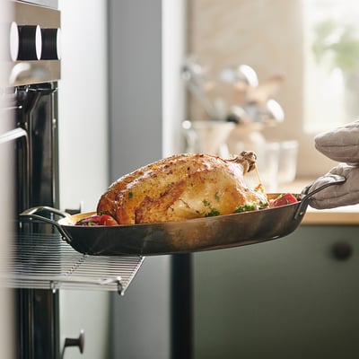 Person holding roasted chicken in black pan in kitchen near oven.