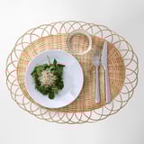 Oval woven rattan place mat with white plate of greens, glass, fork, and knife.