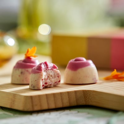 Three pink raspberry cheesecake pralines on a wooden board, one with a bite taken. Topped with edible orange flower petals.