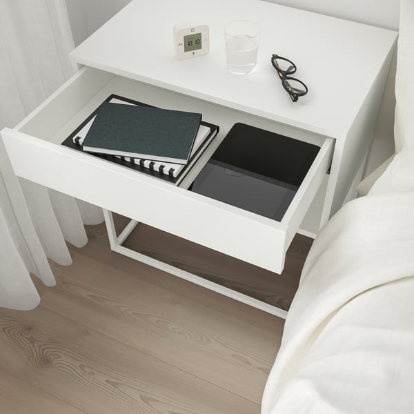 Drawers open silently, showing books and tech, beside a cosy bed. Digital clock and water glass rest atop.