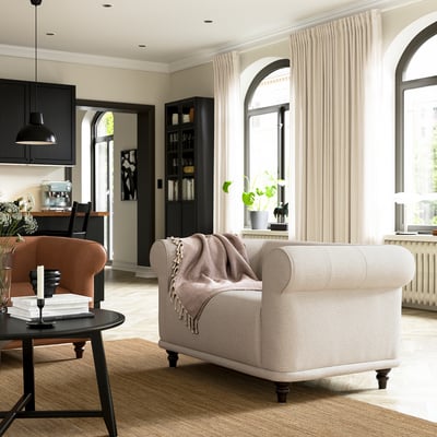 Modern living room: beige armchair, brown velvet chair, black coffee table, large windows with curtains.