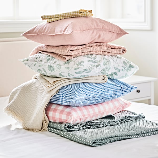 Stack of colourful pillows and blankets: yellow chequered, pink floral, blue chequered, pink gingham, beige, grey. Blue is largest.